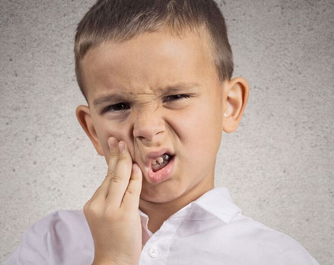 Young boy in white shirt holding cheek, appearing to have tooth pain or discomfort, neutral background.