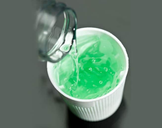Jar pouring green gel or liquid into white cup on gray surface.