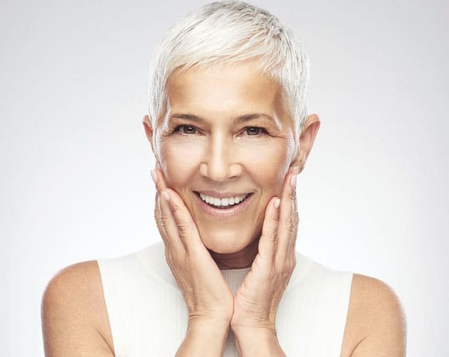 Senior woman with white hair smiling, hands to face, white background studio portrait.