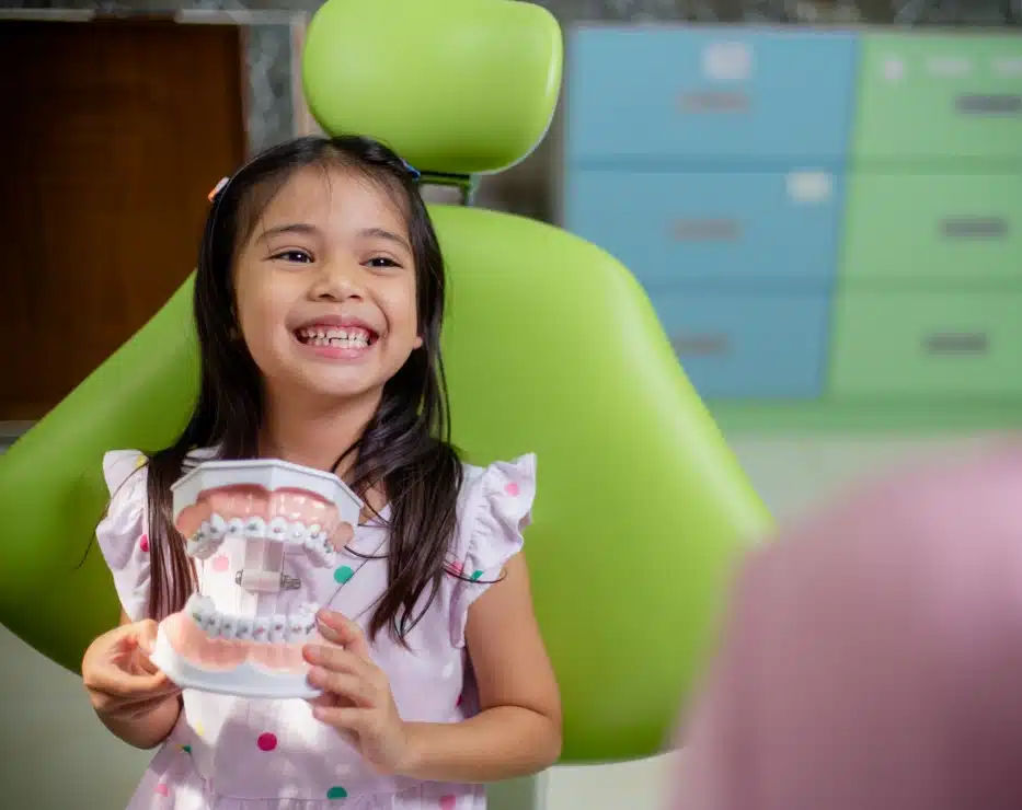 Young girl smiling while holding artwork in front of green chair indoors.