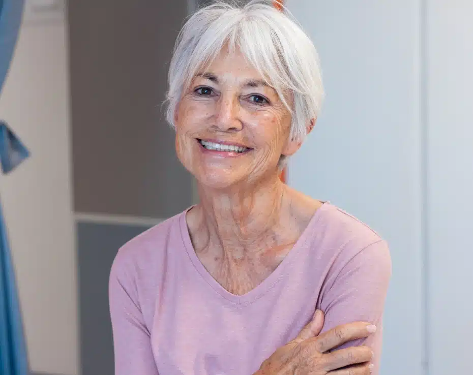 Smiling woman with white hair wearing pink shirt, arms crossed, indoor setting.